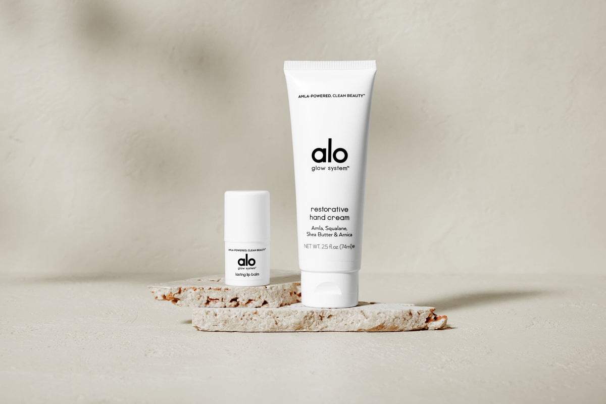 A photo of Alo’s Restorative Hand Cream and Lasting Lip Balm positioned on a tan stone.