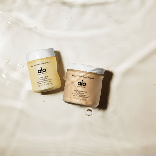 Renew & Glow Body Polish and Mindful Magnesium Bath Soak bottles at the bottom of a beige bathtub under water.