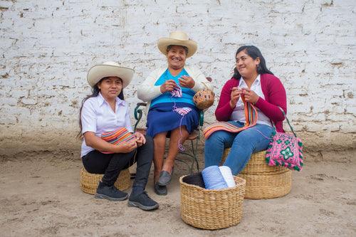 Three women posing in front of a white brick wall and sitting on straw baskets while using an artisanal form of crochet to make bikinis for the Tropic of C x Alo collab.