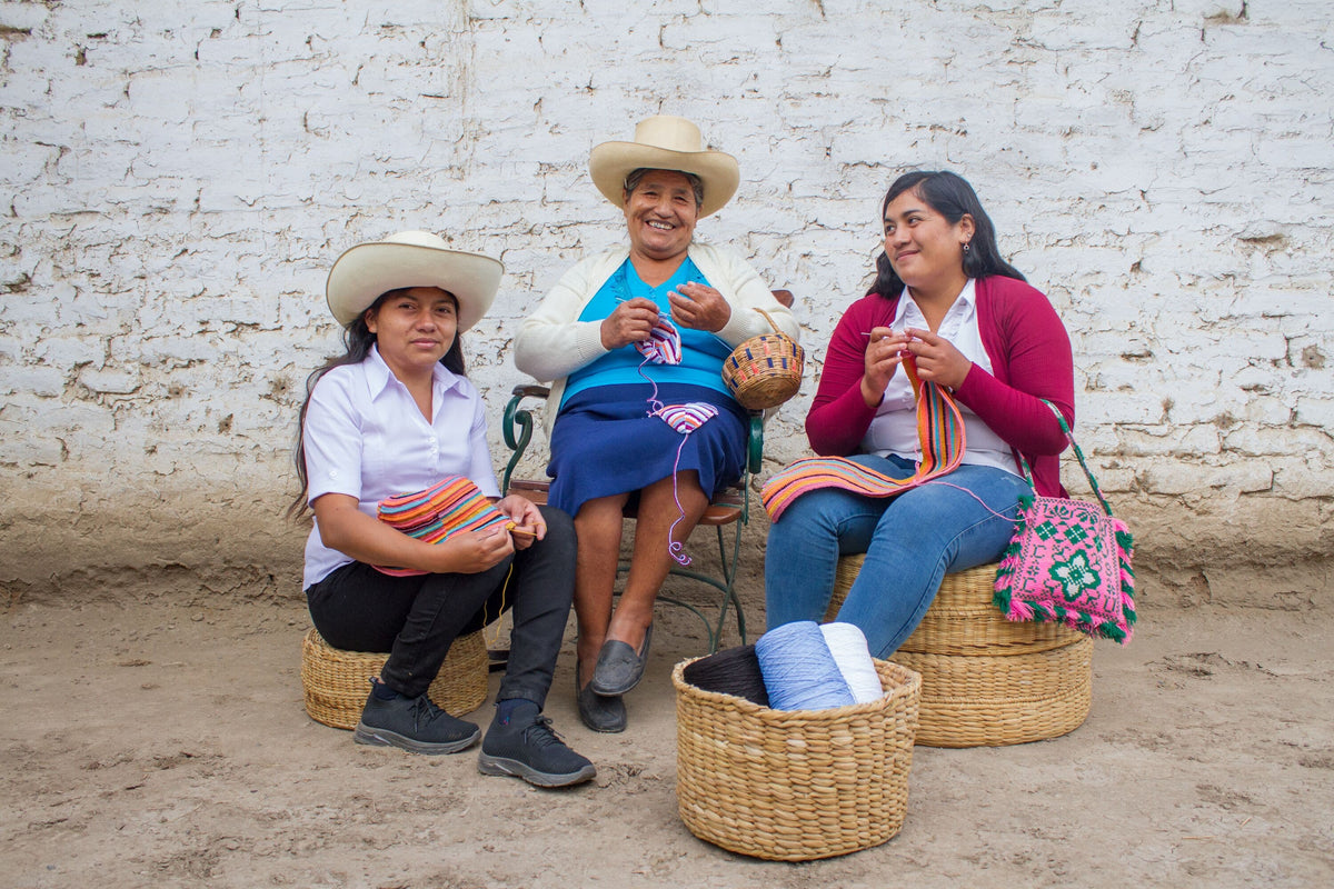 Three women posing in front of a white brick wall and sitting on straw baskets while using an artisanal form of crochet to make bikinis for the Tropic of C x Alo collab.